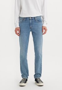 Persoon in lichtblauwe rechte jeans, wit lang mouwen shirt en zwarte lage sneakers met witte zolen en veters.