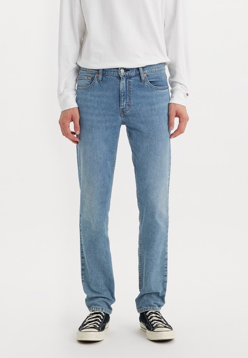 Persoon in lichtblauwe rechte jeans, wit lang mouwen shirt en zwarte lage sneakers met witte zolen en veters.