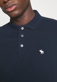 Polo di colore blu navy con pattina a tre bottoni, tessuto testurizzato e un piccolo logo di alce ricamato in bianco sul petto.