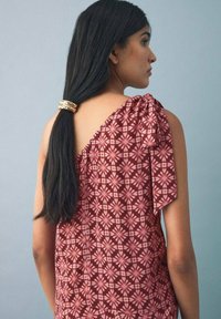 Burgundy topp med vita blommönster, asymmetrisk design och en knuten axeldetalj. Långt mörkt hår som hålls med en gulddetalj.