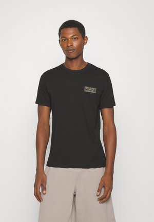 Camiseta de algodón negra con un pequeño logo dorado en el lado izquierdo del pecho, cuello redondo y mangas cortas, combinada con pantalones de color beige claro.