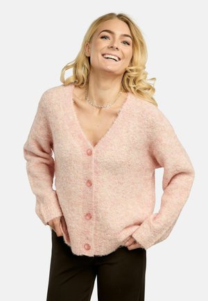 Donna sorridente con capelli biondi ricci che indossa un cardigan rosa peloso con bottoni e pantaloni neri, con le mani poggiate sui fianchi contro uno sfondo semplice.
