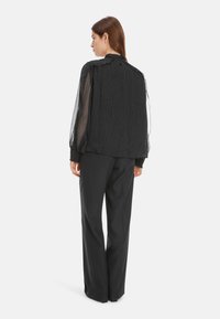 Schwarze gepunktete Bluse mit transparenten Ärmeln, hohem Kragen und lockerer Passform, kombiniert mit maßgeschneiderten schwarzen Hosen. Weicher Stoff und drapierter Schnitt.