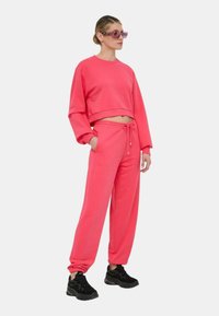 Felpa rosa cropped con maniche lunghe e pantaloni coordinati dotati di vita elastica e tasche laterali, abbinati a sneakers nere.