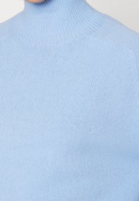 Pull à col roulé bleu clair en tissu tricoté doux avec une texture côtelée. Détails des coutures visibles sur les épaules et le col.