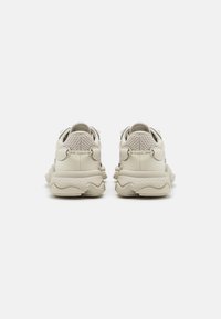 Baskets beige clair avec des panneaux texturés, des lacets ronds et une semelle épaisse et ondulée. Présente un logo discret au niveau du talon et des accents sur le côté.
