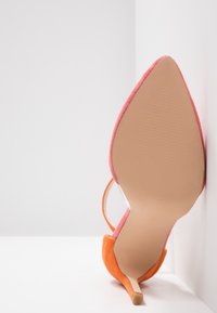 Chaussure à talon haut orange à bout pointu, reposant sur son talon avec la semelle tournée vers l'avant, sur un fond blanc en coin.