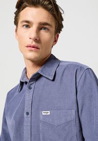 Camisa de pana azul con collar puntiagudo, cierre de botones, bolsillo en el pecho y una pequeña etiqueta con el logo de Wrangler en el bolsillo.