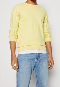 Helles gelbes Langarmshirt mit Rundhalsausschnitt und gerippten Bündchen, kombiniert mit hellblauen Jeans. Der Stoff wirkt weich und dehnbar.