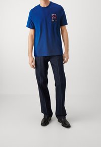 Modré bavlněné tričko s nášivkou na přední straně s vyšívaným písmenem, doplněné tmavými džínami s kontrastním bílým prošitím.