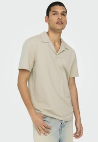Camicia beige a maniche corte con colletto, con un motivo testurizzato, taglio rilassato e scollatura a V. Indossata con jeans in denim azzurro chiaro.