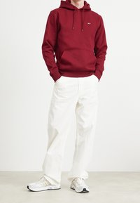 Felpa bordeaux con tasca frontale e coulisse, abbinata a pantaloni bianchi a gamba larga e scarpe da ginnastica leggere con superficie testurizzata.