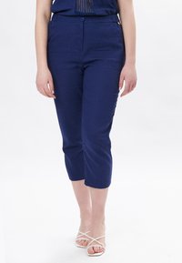 Donkerblauwe cropped broeken van een gestructureerde stof, met een knoopsluiting aan de voorkant en zijzakken, gecombineerd met witte sandalen met bandjes.