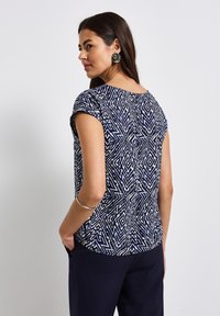 Top a maniche corte con un motivo geometrico blu navy e bianco, scollatura rotonda e vestibilità rilassata. Tessuto liscio con un leggero luccichio.
