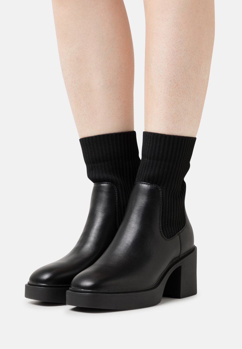 Bottines en cuir noir à talons épais avec des empeignes en maille côtelée style chaussette, portées sur jambes nues sur un fond uni.
