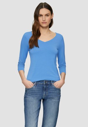 Blauwe, V-hals, lange mouwen top van zachte stof, gecombineerd met lichtblauwe denim jeans. Model staat met handen in de zakken, neutrale achtergrond.