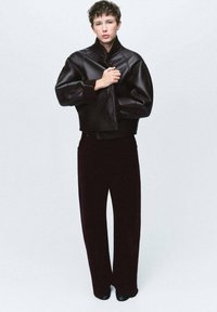 Giovane che indossa una giacca di pelle nera oversize e pantaloni neri larghi, in piedi contro uno sfondo chiaro e uniforme.