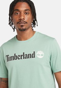 Muž s pletenými vlasy, oblečený v mentolově zeleném tričku Timberland s černým textem a bílým emblémem stromu na bílém pozadí.