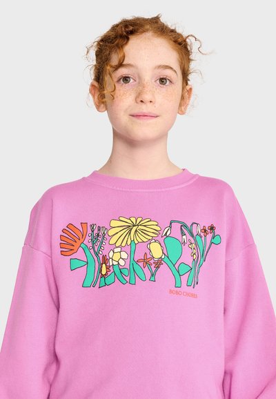 Jeune personne aux cheveux roux bouclés et aux taches de rousseur, portant un sweat-shirt rose avec des illustrations florales colorées sur la poitrine.