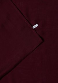 Tissu bordeaux foncé avec une petite étiquette blanche rectangulaire portant l'inscription "CECIL" cousue dans une couture.