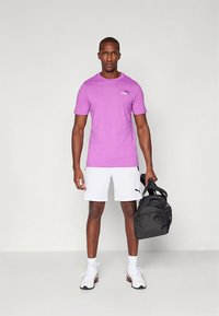 Pánske fialové tričko s krátkym rukávom s logom Puma, v kombinácii s bielymi šortkami a čiernou športovou taškou, stojace na neutrálom pozadí.