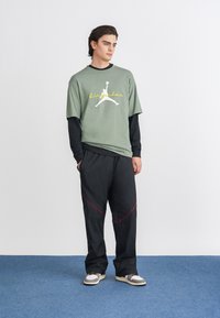 Πράσινο oversized t-shirt με λευκό και κίτρινο γραφικό Air Jordan, σε συνδυασμό με μαύρο παντελόνι με κόκκινες λεπτομέρειες και άνετη εφαρμογή.