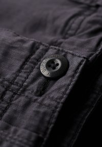 Zwarte knoop met vier gaten, gegoten met "SUPERDRY," op donkere stof met subtiele textuur en stiksels.