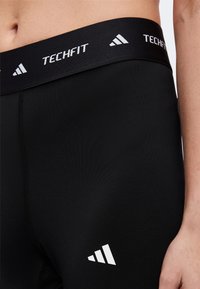 Gros plan d'une personne portant un short de compression Adidas Techfit noir avec logo sur la ceinture et la cuisse.