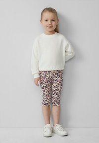 Biały sweter, wzorzyste beżowe legginsy w różowe serca i białe sneakersy. Dziecko stoi z jedną ręką na biodrze, uśmiechając się.