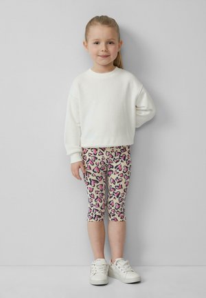 Weißer Sweatshirt, gemusterte beige Leggings mit pinken Herzen und weiße Sneaker. Kind steht mit einer Hand an der Hüfte und lächelt.