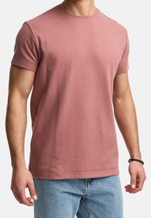 Man wearing a plain dusty pink short-sleeve t-shirt and light blue jeans, with a black braided bracelet on his right wrist.