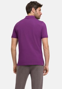 Polo shirt violet à manches courtes, fabriqué en tissu lisse. Il dispose d'un col classique et d'ourlets fendus sur les côtés, porté avec un pantalon gris.