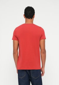 Červené tričko s krátkým rukávem, lehký bavlněný materiál, kulatý výstřih, těsný střih, hladká textura, nošeno s modrými džínovými kalhotami, pohled zezadu.