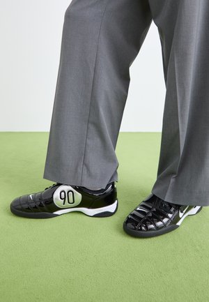 Baskets en cuir verni noir avec un détail argenté "90", présentant une surface texturée et un design à lacets, associées à un pantalon large gris.