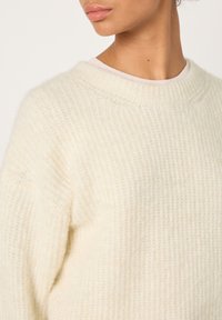 Elnių megztinis dramblio kaulo spalvos, su struktūruotu ribuotu paviršiumi, plačia valtine apykakle ir laisvo silueto, pasižymi subtiliomis siuvimo detalėmis ir minkštu išvaizdu.