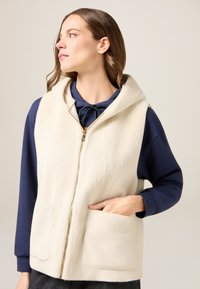 Gilet in pile color crema con cappuccio, dotato di cerniera frontale e due tasche laterali. Indossato sopra una maglia a maniche lunghe blu navy. Tessuto morbido.