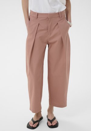 Személy magas derekú, széles szárú, piszkos rózsaszín árnyalatú, elején redőzött nadrágot visel, fehér felsővel, fekete papucsban és ezüst órával a bal csuklóján.