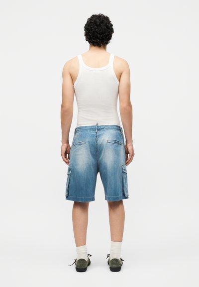 Άνδρας με σγουρά μαλλιά που φοράει λευκό φανελάκι, τζιν cargo σορτς, λευκές κάλτσες και σκούρα αθλητικά παπούτσια, στέκεται γυρισμένος με την πλάτη σε λευκό φόντο.