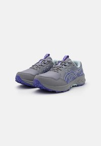 Sapatos atléticos cinzentos com detalhes em roxo, parte superior em malha e sintético, sola texturizada e sistema de atacadores. Apresenta uma aba de puxar no calcanhar.