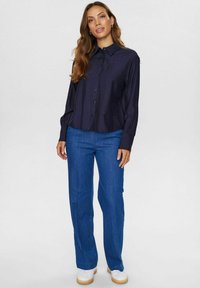 Marineblå knap-rigtig bluse med en let blank tekstur, parret med lige-skårede blå bukser og hvide sneakers. Blusen har samlet ærmer.