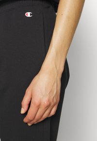 Pantalones jogger negros con una textura suave, con una cintura elástica y un pequeño logo rojo y blanco en el muslo, primer plano de una mano descansando sobre la tela.