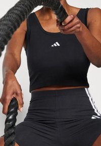 Czarny, krótki sportowy top z białym logo Adidas, w zestawie z czarnymi szortami o wysokim stanie z białymi paskami po bokach, trzymając liny do walki.