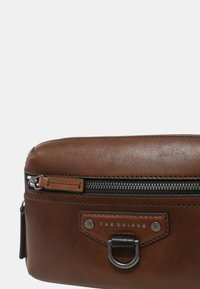 Borsa in pelle marrone con una superficie liscia, una tasca frontale con zip e un anello in metallo. Logo "THE BRIDGE" impresso sul davanti.