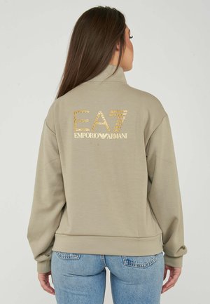 Donna che indossa una felpa beige con il logo dorato "EA7 Emporio Armani" sulla schiena, abbinata a jeans azzurri, di spalle su sfondo semplice.