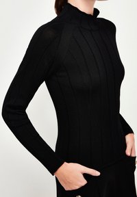 Woman wearing a fitted black ribbed long-sleeve top with a high neck, one hand in pants pocket, against a plain light background.