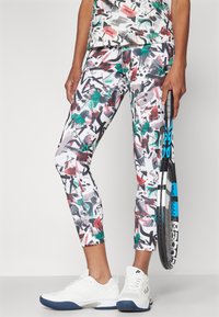 Eng anliegende, cropped Leggings mit einem mehrfarbigen, abstrakten Muster in Schwarz, Grün und Rot. Kombiniert mit weißen Sportschuhen und einem Schläger.