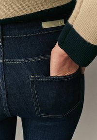 Jeans de mezclilla oscura con costuras visibles, un bolsillo trasero y una etiqueta de marca. Parcialmente cubiertos por un suéter de punto con colores contrastantes.