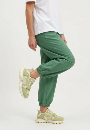 Person trägt grüne Jogginghose, weiße und gelbe Sneakers und ein weißes kurzärmeliges Shirt, steht auf einem Bein vor einfachem Hintergrund.