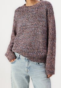 Pull en maille mêlant des fils violets, marron et bleus ; coupe décontractée avec manches longues et poignets côtelés ; porté sur un jean clair.