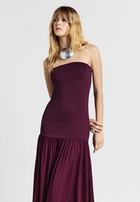 Strapless burgundy dress with a fitted bodice and flowing, gathered skirt. Silver pendant necklace adds contrast to the design. Smooth texture.